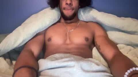 Media: Video of a light-skinned man with a beard and medium-length black curly hair, shirtless under a light blue blanket, lying on a bed with white sheets, wearing a silver necklace.