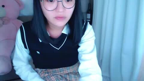 Media: Video of a young Asian woman with shoulder-length black hair, wearing glasses, a black sweater vest over a white collared shirt, and a plaid skirt, sitting indoors near a stuffed bear.