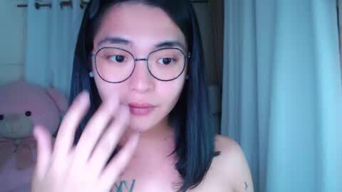Media: Video of an Asian woman with long black hair, glasses, and a pink tattoo on her chest, applying lipstick in a bathroom with light-colored walls and a stuffed bear.